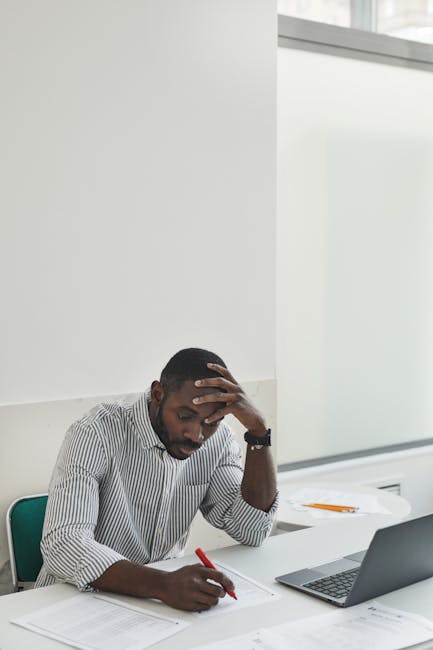 Person sitting at a desk with a laptop, looking thoughtful and slightly tired.